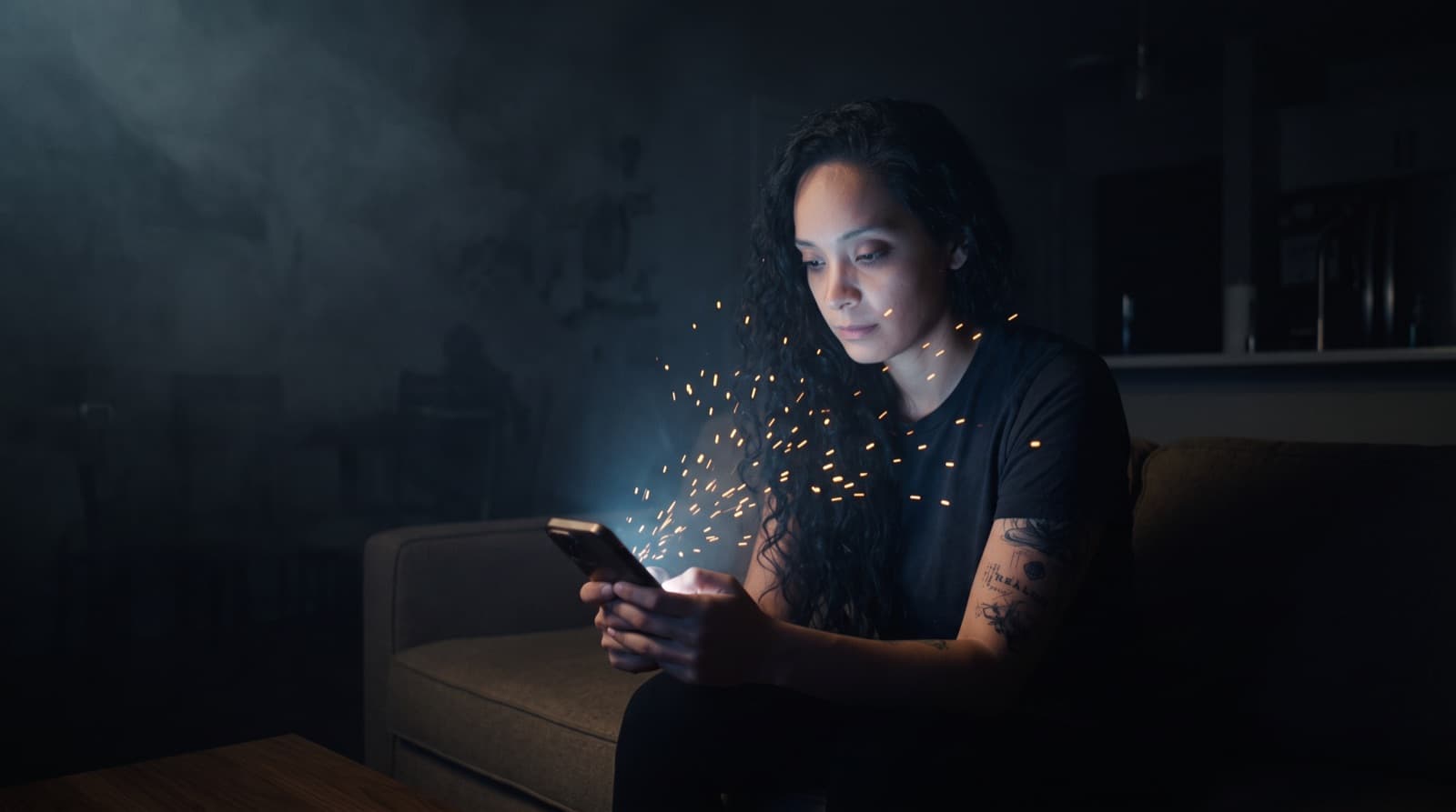 Mel Cobian on a couch in a dark room, golden sparks streaming from her into the glow of her phone