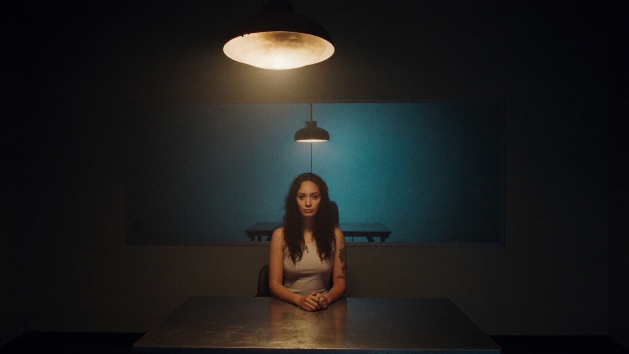 Mel Cobian seated alone at a table under a single hanging lamp, a two-way mirror behind her