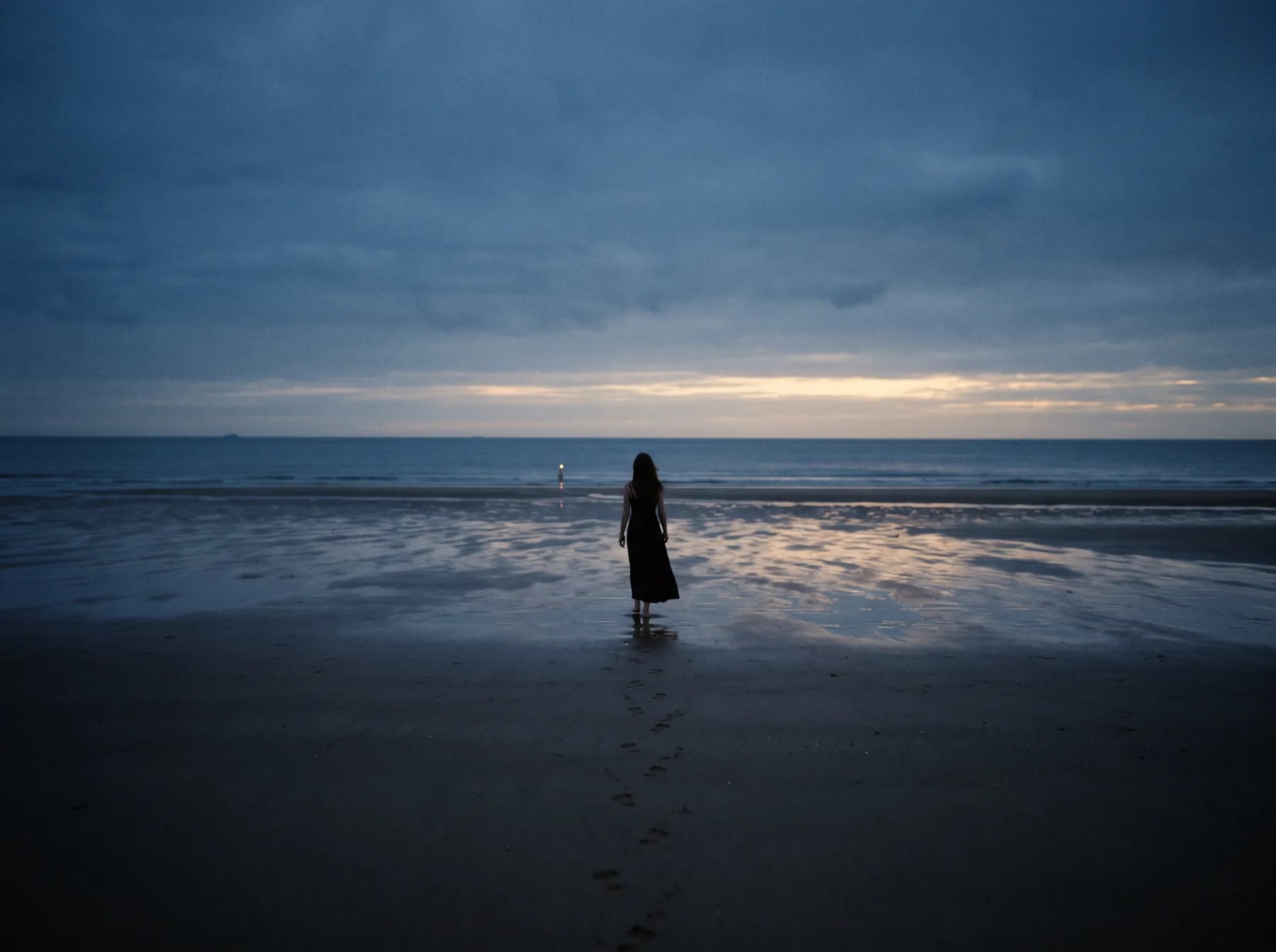 A figure standing at the water's edge at dusk, footprints trailing behind in the sand