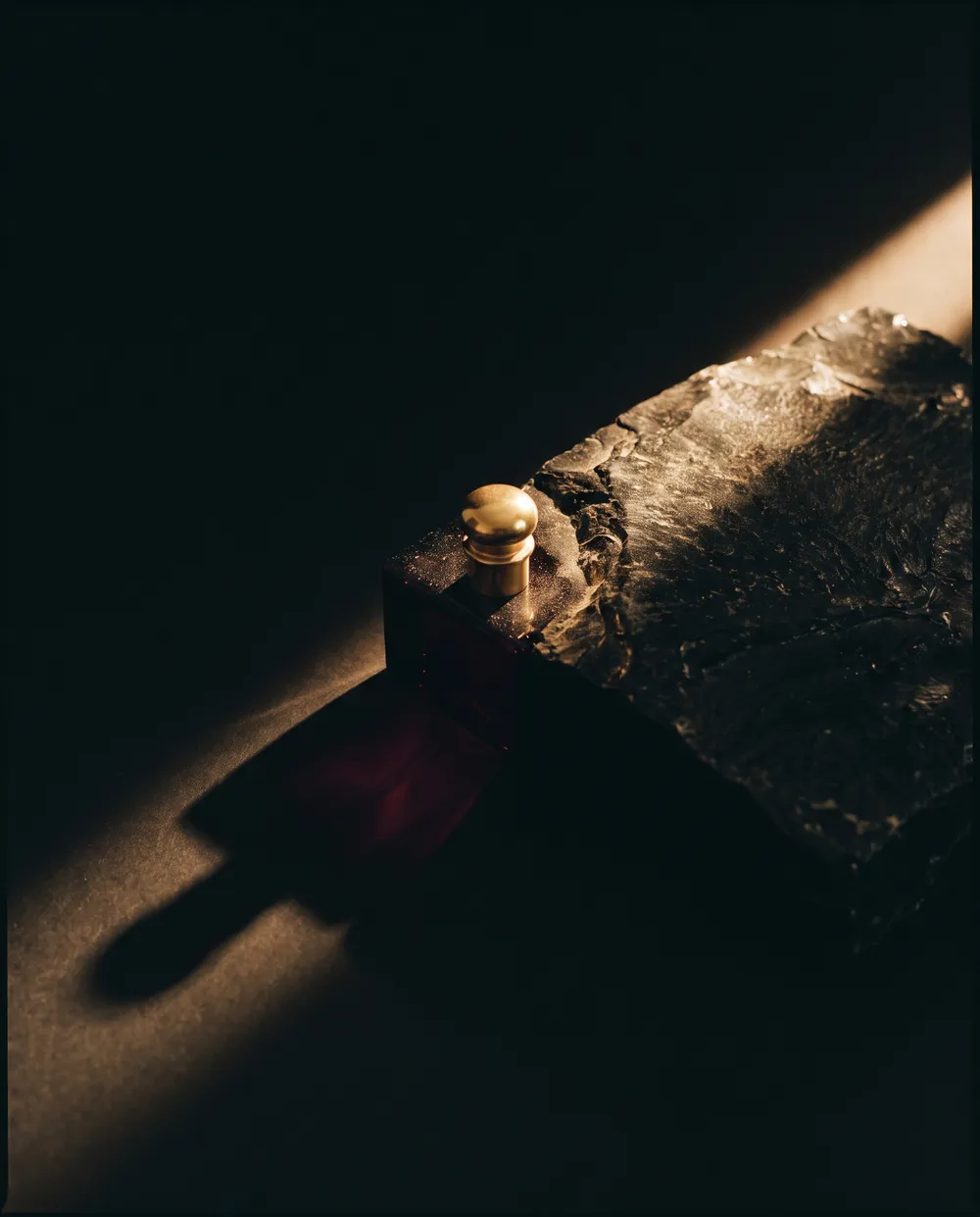 A shaft of gold light across dark textured stone, one small polished object casting a long shadow