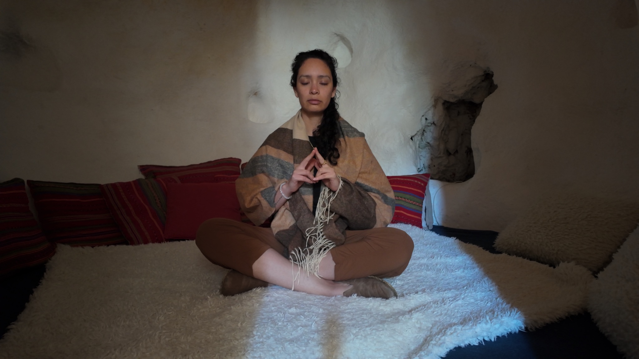 Mel Cobian seated cross-legged in a stone room, wrapped in a woven blanket, hands joined and eyes closed